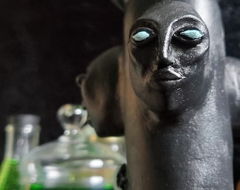 May include: A black and gray ceramic sculpture of a face with blue eyes. The sculpture is on a wooden surface with a glass jar and other objects in the background.