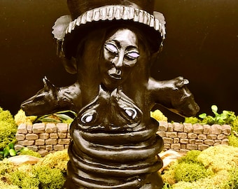 May include: Black ceramic sculpture of a woman with a top hat, wearing a snake and holding two animal heads. The sculpture is on a bed of green moss.