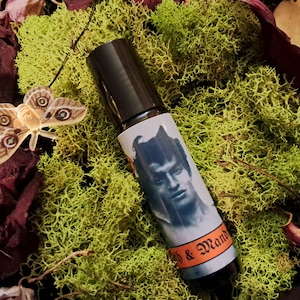 Pode incluir: Um frasco preto com rolo com um rótulo branco que apresenta uma ilustração em preto e branco de um homem com chifres. O rótulo também inclui o texto "Witch & Wand".