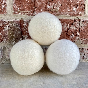 May include: Three white wool dryer balls. The balls are round and fluffy, and they are arranged in a triangle shape.
