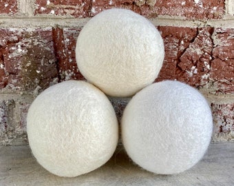 May include: Three white wool dryer balls. The balls are round and fluffy, and they are arranged in a triangle shape.