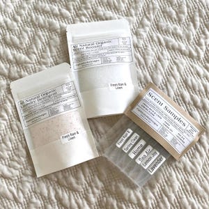 May include: Three white paper bags with brown labels containing laundry detergent and scent booster samples. The labels have the text "All Natural Organic Laundry Detergent", "All Natural Organic Scent Booster", and "Scent Samples". The scent samples include "Fresh Rain & Linen", "Spring Clean", "Home", "Sea Air", "Lemon Joy", and "Lavender".