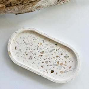 May include: An oval-shaped decorative tray with a speckled terrazzo design. The tray features a cream-colored base with embedded chips of various colors, including brown, gray, and white. A piece of driftwood rests in the background.