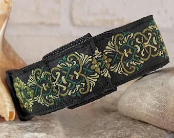 May include: A black fabric strap featuring an intricate embroidered pattern in shades of olive green, forest green, and gold. The strap has a rectangular shape with a visible hook-and-loop fastener closure on the front surface of the material.