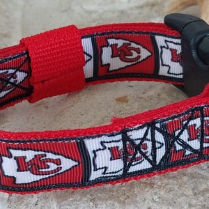 May include: Red, white and black dog collar with a repeating pattern of the Kansas City Chiefs logo. The collar has a black plastic buckle and a silver D-ring.