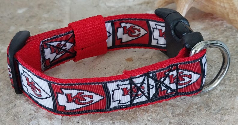 May include: Red, white and black dog collar with a repeating pattern of the Kansas City Chiefs logo. The collar has a black plastic buckle and a silver D-ring.