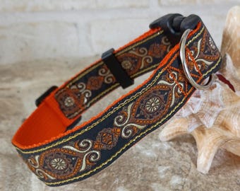 May include: A pet collar featuring a black fabric strap with an intricate woven pattern in shades of orange, brown, and gold. The collar includes a bright orange inner lining, a black plastic side-release buckle, and a silver metal D-ring for a leash.