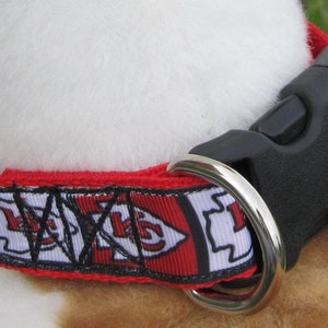 May include: Red, white and black dog collar with a black plastic buckle and a silver metal ring. The collar has a repeating pattern of a red arrow with a white outline and the letters "KC" in black.