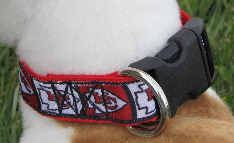 May include: Red, white and black dog collar with a black plastic buckle and a silver metal ring. The collar has a repeating pattern of a red arrow with a white outline and the letters "KC" in black.
