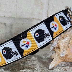 Op de afbeelding: Een zwart, wit en geel halsbandje voor huisdieren met een herhalend patroon van footballhelmen met een cirkelvormig logo. De halsband is gemaakt van geribbelde stof en heeft aan één uiteinde een zilverkleurige metalen D-ring.