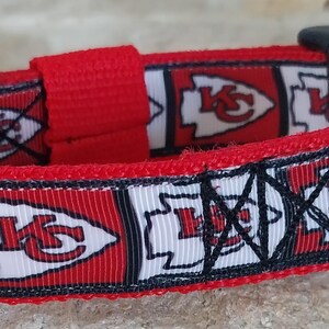 May include: Red and white dog collar with a repeating pattern of the Kansas City Chiefs logo. The collar has a black buckle and a silver D-ring.