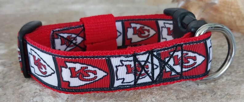 May include: Red and white dog collar with a repeating pattern of the Kansas City Chiefs logo. The collar has a black buckle and a silver D-ring.