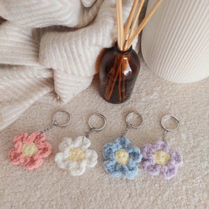 Könnte beinhalten: Vier handgefertigte Schlüsselanhänger in Blumenform in Pastellfarben: Rosa, Weiß, Blau und Lila. Jede Blume hat eine gelbe Mitte und einen silbernen Schlüsselring. Die Schlüsselanhänger sind aus einem weichen, strukturierten Material.