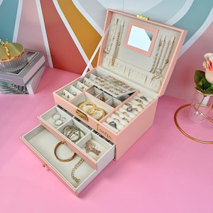 May include: A pink jewelry box with two drawers and a mirror. The top drawer has compartments for earrings, rings, and other small jewelry. The bottom drawer has a larger compartment for necklaces and bracelets. The box is personalized with the name "Laura".