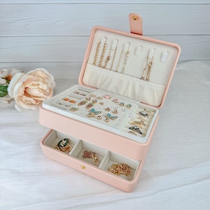May include: A two-tiered pink jewelry box with a hinged lid. The top tier has a white felt lining with slots for necklaces and bracelets. The bottom tier has a drawer with compartments for rings and earrings. The box is filled with various jewelry, including earrings, rings, necklaces, and bracelets.