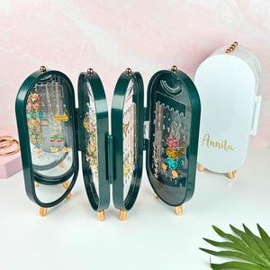 May include: A green jewelry organizer with a mirrored door and multiple compartments for storing earrings, necklaces, and bracelets. The organizer has a gold-colored base and is open to reveal the jewelry inside. The name "Annita" is written on a white jewelry organizer in the background.