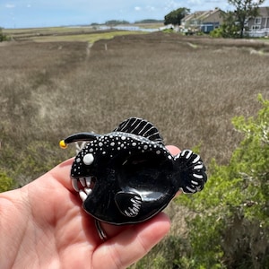 May include: A black anglerfish figurine with white spots and sharp teeth. The figurine has a yellow lure and white accents. The fish is held in a hand against a backdrop of a marsh and blue sky.