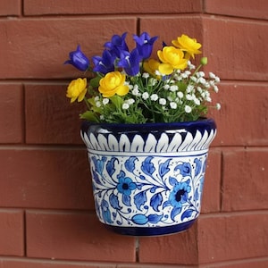 Puede incluir: Una maceta de pared de cerámica azul y blanca con un diseño floral, llena de flores amarillas, azules y blancas. La maceta está montada en una pared de ladrillo rojo, mostrando una exhibición decorativa y colorida.