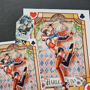 May include: Illustration prints featuring Harley Quinn in a playing card design. The artwork showcases Harley Quinn in a dynamic pose, with a diamond pattern background. The prints include the text "Harley Quinn" and the artist's logo.