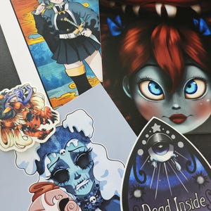 May include: A collection of stickers featuring various illustrations. One sticker features a blue and white illustration of a creature with a large mouth and eyes. Another sticker features a blue and white illustration of a creature with a large eye and a teardrop. The text "Dead Inside" is printed on the sticker.