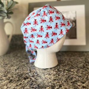 May include: A blue and red patterned surgical scrub hat with a lobster design. The hat has a tie on the back.