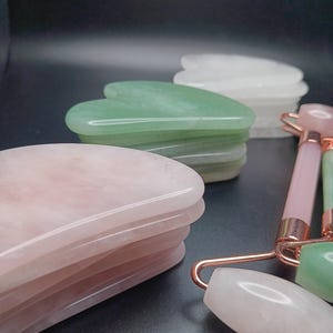 May include: A collection of facial massage tools, including rose quartz and jade gua sha stones and facial rollers. The gua sha stones are stacked in pink, green, and white. The rollers have pink and green handles with rose gold accents.