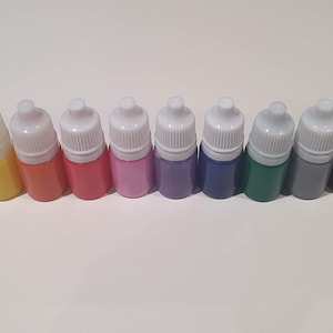 Peut inclure: Un ensemble de 12 petits flacons de peinture, chacun avec un bouchon compte-gouttes blanc. Les flacons sont disposés en rangée de gauche à droite, avec les couleurs blanc, jaune, orange, rouge, rose, violet, bleu, vert, gris, marron, noir et bleu.