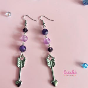 May include: A pair of silver arrow earrings with purple and lavender beads. The earrings are on a pink background. leishi made with love.