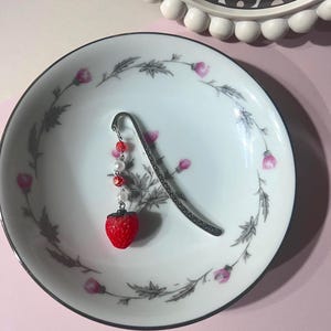 May include: A silver bookmark with a red and white strawberry charm and white beads hanging from it. The bookmark is on a white plate with a floral design.