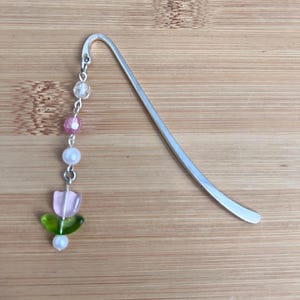 May include: A silver bookmark with a pink and green flower charm. The charm is made of beads and hangs from the bookmark with a silver chain.
