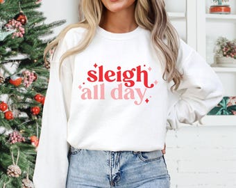 Könnte beinhalten: Ein weißes Langarm-Sweatshirt mit Rundhalsausschnitt und dem roten sowie rosafarbenen Schriftzug "sleigh all day" mittig auf der Brust. Die Grafik enthält kleine vierzackige Sternakzente um den Text, der in einer fetten Schriftart gedruckt ist.
