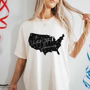 May include: A cream-coloured t-shirt with a black silhouette of the United States map filled with stars. The text "The United States of America" is written in a script font within the map. The shirt has short sleeves.