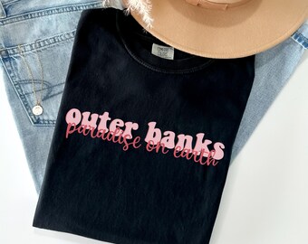 May include: A black short-sleeved t-shirt featuring the text "outer banks" in large, light pink bubble letters and "paradise on earth" in a smaller, cursive red font. The shirt is layered with blue denim jeans, a tan hat, and a gold necklace.