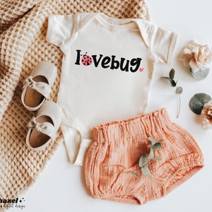 Puede incluir: Un body blanco con el texto "I lovebug" en negro con un gráfico de mariquita rosa. El body se combina con un short rosa con volantes.