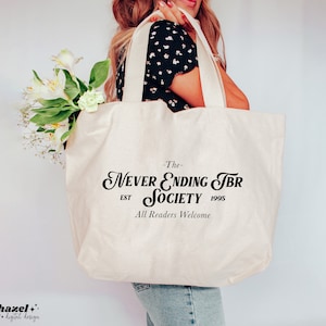 K&ouml;nnte beinhalten: Eine wei&szlig;e Canvas-Tragetasche mit schwarzem Text, der "The Never Ending TBR Society Est 1995 All Readers Welcome" lautet.