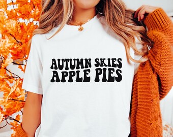 May include: White t-shirt with black text that reads "Autumn Skies Apple Pies" in a retro font.