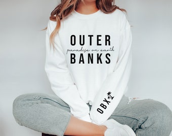 Può includere: Felpa bianca con testo nero che recita "Outer Banks paradise on earth". La manica sinistra ha un testo nero che recita "OBX" con una grafica di palma.