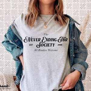 K&ouml;nnte beinhalten: Graues T-Shirt mit dem Text "The Never Ending TBR Society Est 1995 All Readers Welcome" in Schwarz.
