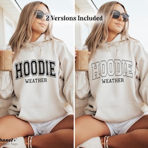 Op de afbeelding: Een beige hoodie met de tekst "Hoodie Weather" in zwarte blokletters. De hoodie wordt gedragen door een persoon die een kop koffie vasthoudt.