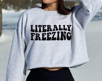 以下が含まれることがあります： 胸元に波打つレトロなフォントで黒い文字「LITERALLY FREEZING」がプリントされた、ライトグレーの長袖クルーネックトレーナー。リブ編みの袖口と襟元を備えた、ゆったりとしたシルエットのカジュアルなトップスです。