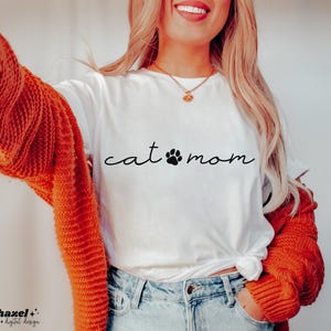 May include: A white short-sleeved t-shirt featuring the black text "cat mom" with a paw print replacing the letter "o". The shirt is worn under a chunky, long-sleeved, burnt orange knit cardigan and paired with light blue denim jeans.