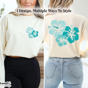 May include: A white long-sleeved shirt with a design of three teal hibiscus flowers on the back. The shirt is shown being worn by a person with black pants and blue jeans.
