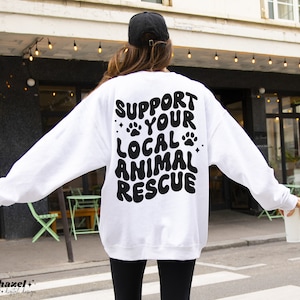 May include: White sweatshirt with black text that reads "Support Your Local Animal Rescue" in a retro, wavy font. The text is surrounded by paw prints and stars.