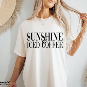 Könnte beinhalten: Cremefarbenes T-Shirt mit dem Text "SUNSHINE & ICED COFFEE" in fetter, schwarzer Schrift. Das Shirt hat kurze Ärmel und eine lockere Passform. Das Design ist mittig auf der Vorderseite platziert. Der Markenname "hazel + digital design" befindet sich unten links.