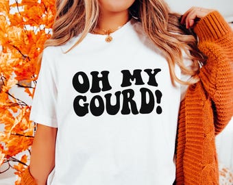 May include: A white short-sleeved t-shirt featuring the black text OH MY GOURD! printed in a bold, rounded font across the chest. The shirt is worn under a thick, textured orange knit cardigan, showcasing a simple round neckline and short sleeves.