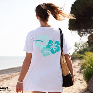 May include: White t-shirt with a blue and green hibiscus flower design on the back. The shirt is being worn by a person walking on a beach.