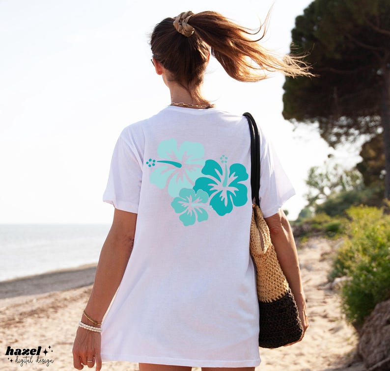 May include: White t-shirt with a blue and green hibiscus flower design on the back. The shirt is being worn by a person walking on a beach.