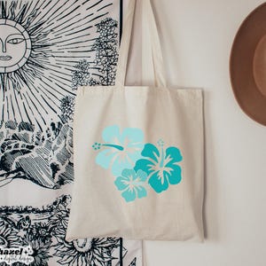 May include: A white canvas tote bag with a design of three blue and turquoise hibiscus flowers. The bag has long straps and is perfect for carrying everyday essentials.