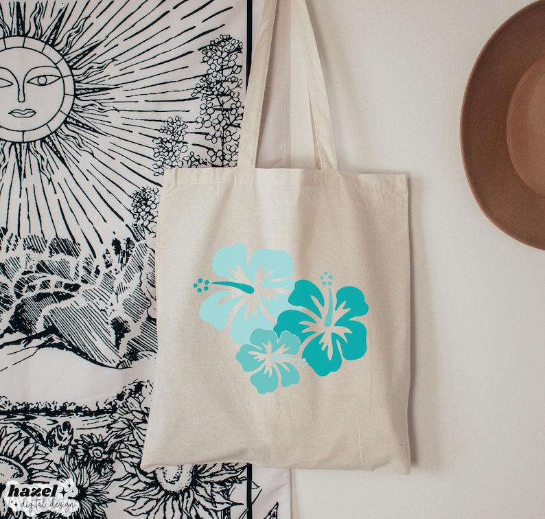 May include: A white canvas tote bag with a design of three blue and turquoise hibiscus flowers. The bag has long straps and is perfect for carrying everyday essentials.