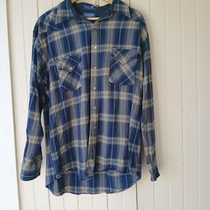 May include: Blue and tan plaid button-down shirt with two chest pockets. The long-sleeved shirt is hanging on a wooden hanger. The shirt has a classic plaid pattern in shades of blue and tan.